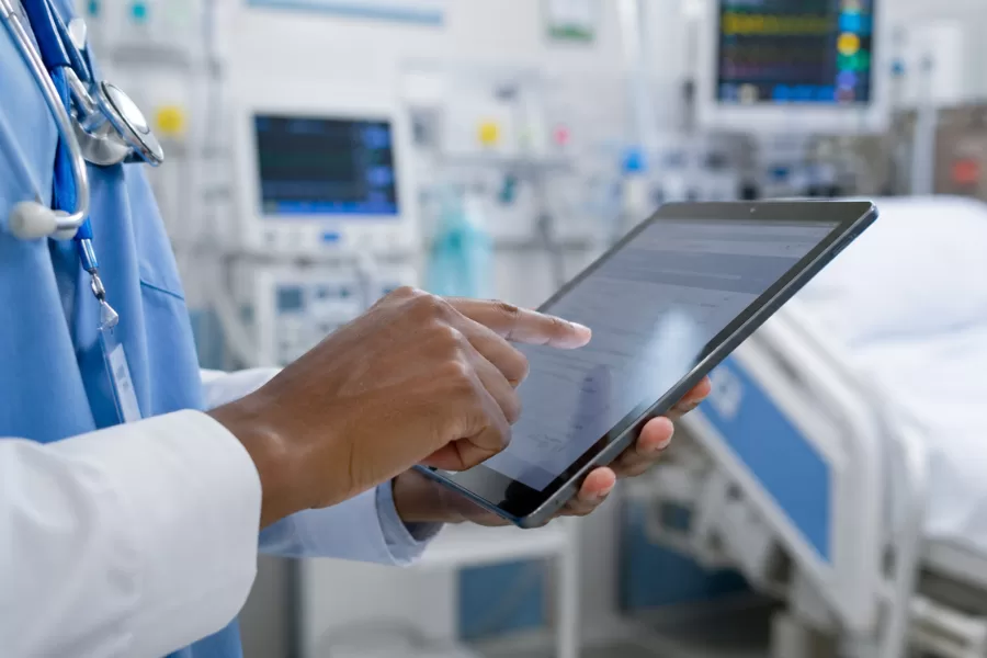 Carreira Hospitalar na Enfermagem: Guia Prático para Começar e Evoluir Profissional de enfermagem usando tablet para registro de prontuário eletrônico ao lado de leito hospitalar