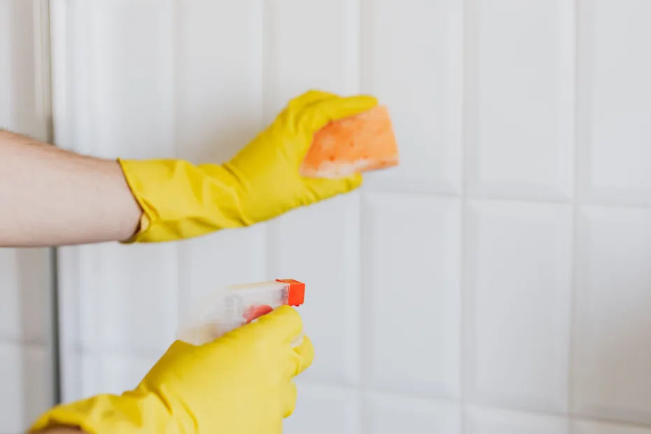 Manos con guantes limpiando azulejos blancos con esponja y producto de limpieza en un baño