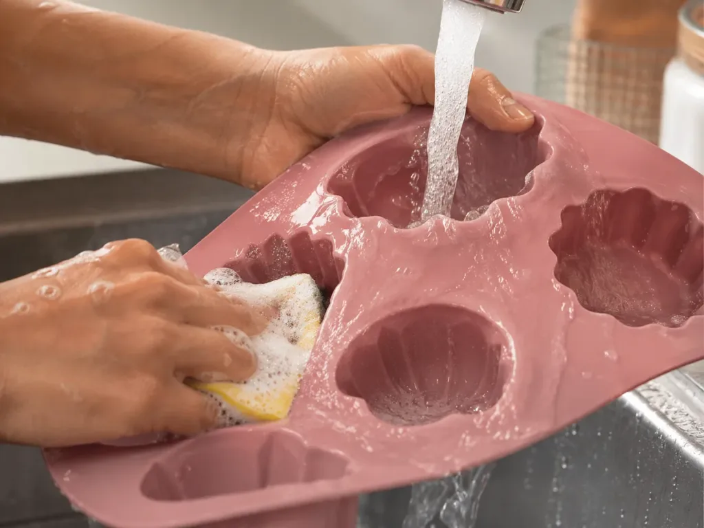 Manos lavando y enjuagando un molde de silicona con esponja suave bajo agua corriente